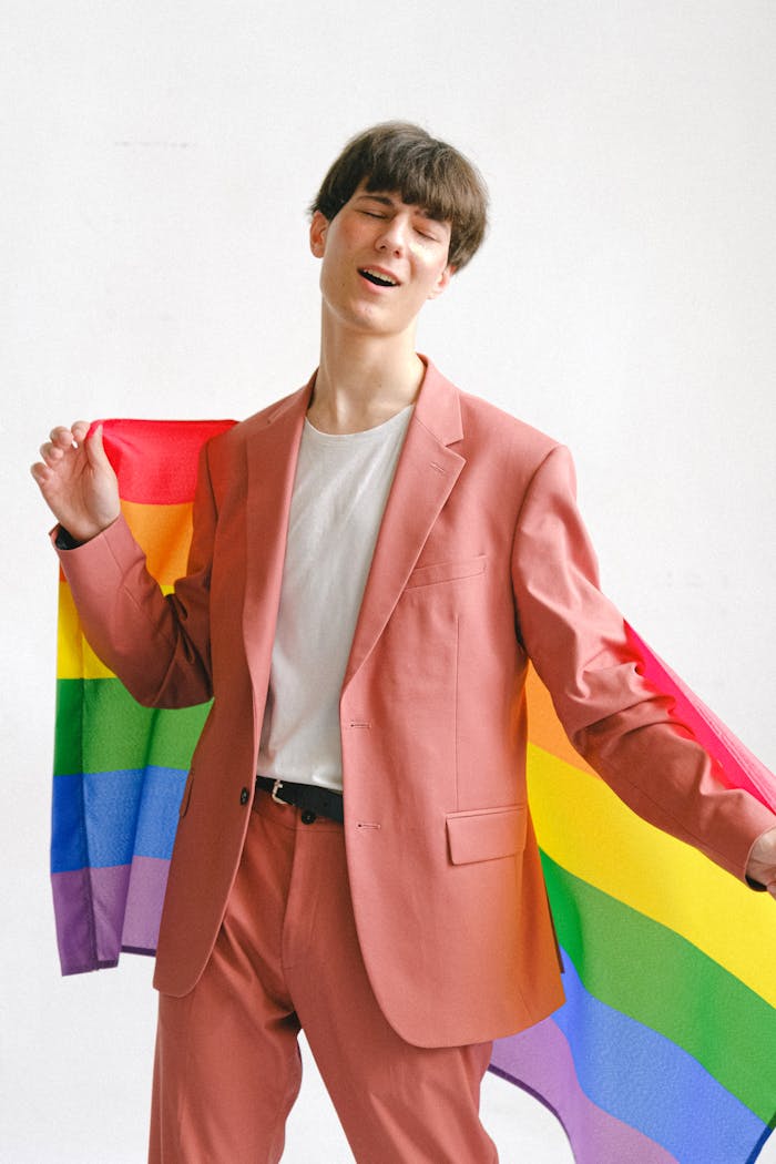 A young man in a pink suit joyfully holding a rainbow LGBTQ pride flag indoors.